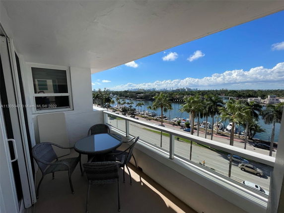 Balcony view overlooking a waterfront and a road, with outdoor seating and a cityscape in the distance.