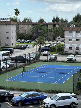 View of a fenced tennis court near residential buildings and parking area.