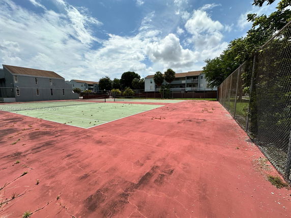 View of a tennis court surrounded by chain-link fencing with buildings in the background.