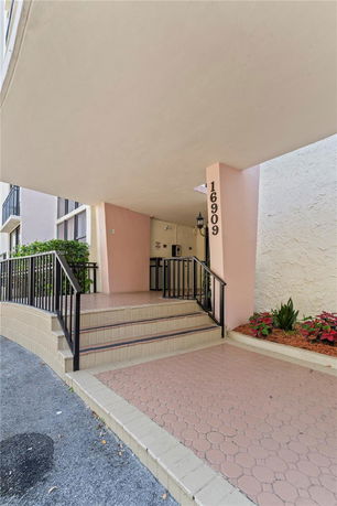 Front entrance of a building with a staircase leading to the entrance, featuring a pink wall with the number 16909 displayed.