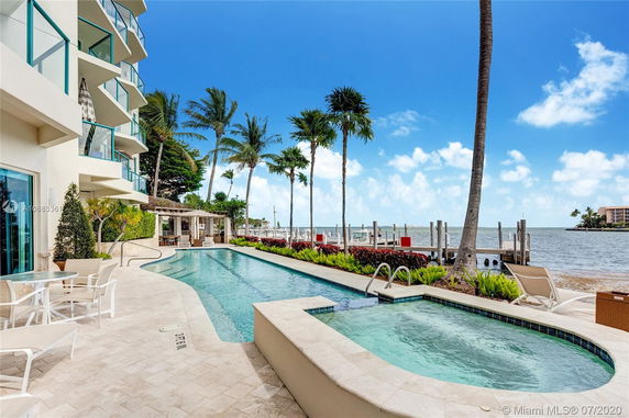 View of an outdoor pool area adjacent to a waterfront, featuring a multi-story building with balconies and tropical landscaping.