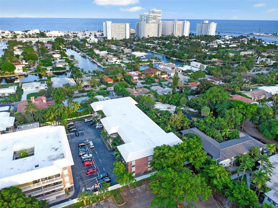 Aerial view of residential and commercial buildings near the coastline with greenery and parking areas visible.