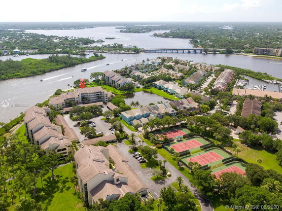 Aerial view of residential complex near waterway with multiple buildings, roads, and tennis courts.