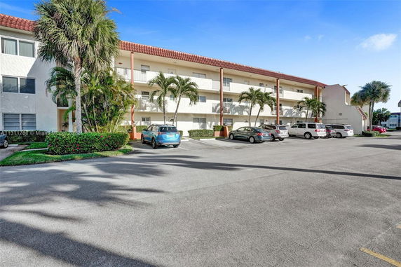 Parking area in front of a multi-level building with balconies and palm trees.