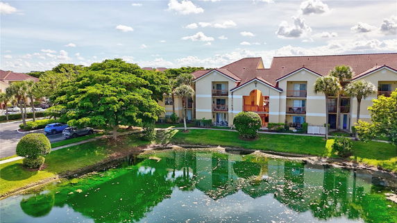 Front view of a multi-story building with balconies and a gabled roof, set near a pond with clear reflections.