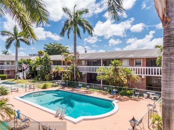 Outdoor swimming pool area with a two-story building in the background, surrounded by palm trees.