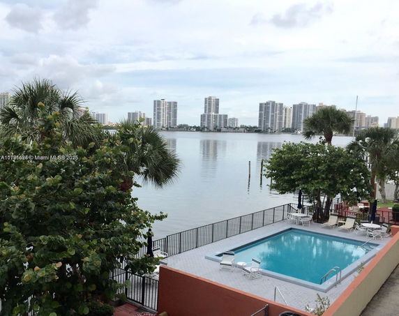 View of a swimming pool with water in the background and high-rise buildings across the water.