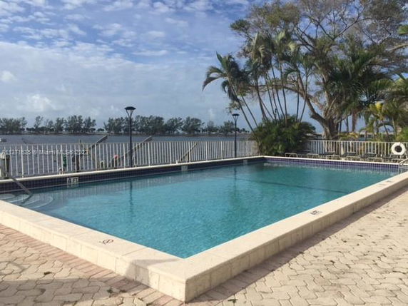 Swimming pool area with view of a waterfront and trees in the background.
