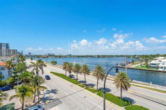 Panoramic view of a coastal road with palm trees, buildings, and a body of water.