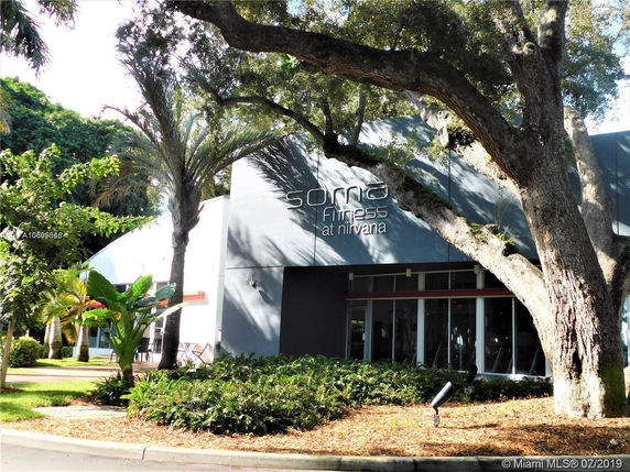 Front view of a fitness center with large windows, surrounded by trees and greenery.