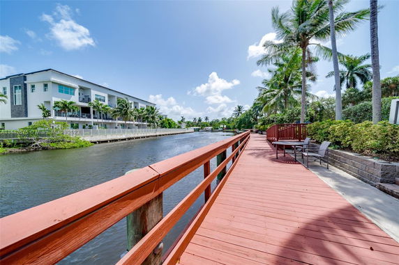 View of a modern multi-story building next to a canal with a boardwalk and palm trees.
