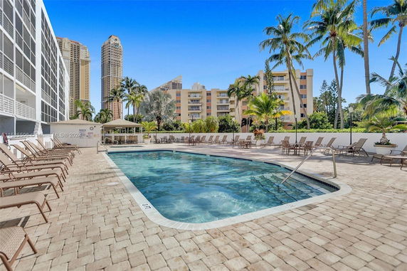 Outdoor swimming pool area with lounge chairs, surrounded by palm trees and buildings.