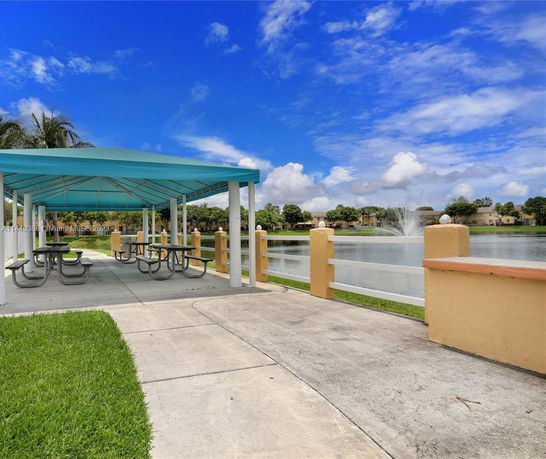 Outdoor seating area with picnic tables and a blue canopy by a lake with a fountain.