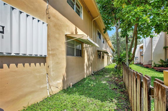 Side view of a beige building with windows and small awnings, alongside a grassy area and a wooden fence.