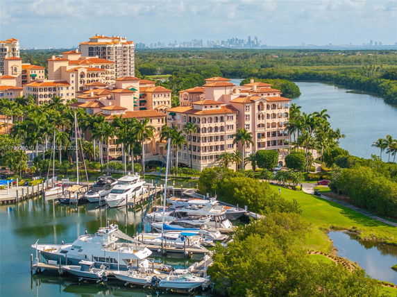 Aerial view of an apartment complex by a marina with boats docked, surrounded by lush greenery and water, with a city skyline in the background.