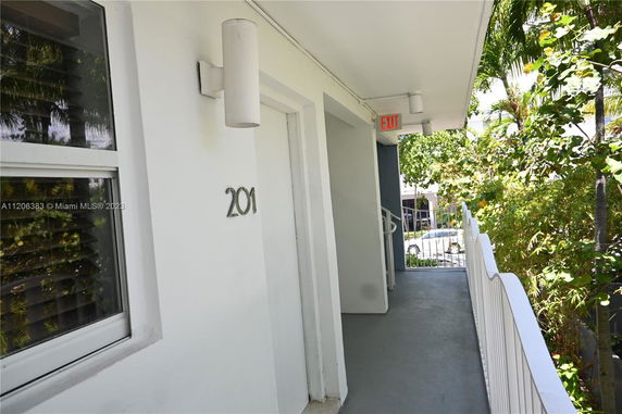 Outdoor view of a building corridor with a door labeled 201, featuring greenery in the surroundings.