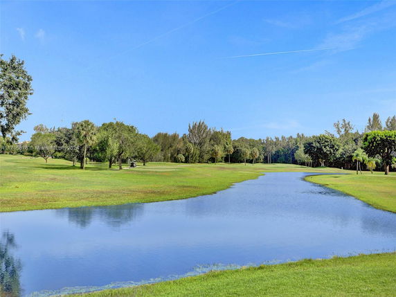 Scenic view of a grassy landscape with a pond and trees under a blue sky.