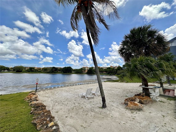 View of a sandy beach area with palm trees and a body of water in the background.
