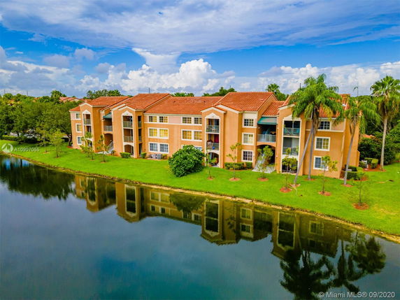 Front view of a multi-story apartment building with a red roof and balconies, located next to a water body.