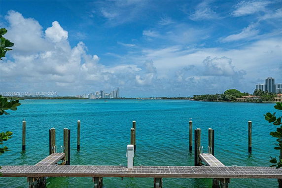 View of a waterfront area with a dock extending into a bright blue body of water, with distant buildings and greenery on the horizon.