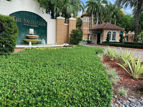 Front view of a building entrance with a sign and fountain, surrounded by greenery and palm trees.
