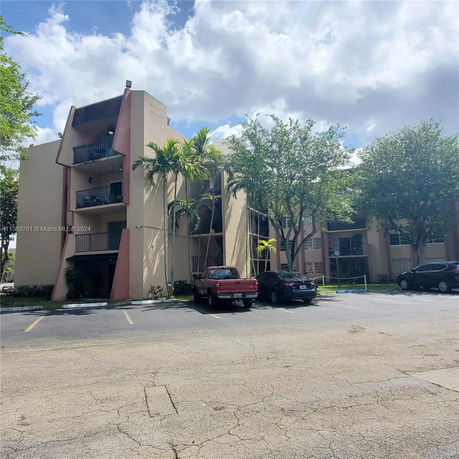 Front view of a multi-story residential building with balconies and adjacent parking area.