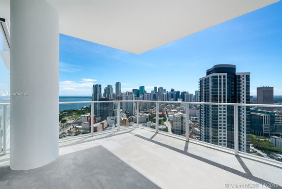 A high-rise apartment balcony with a glass railing offering a view of a city skyline and water body.