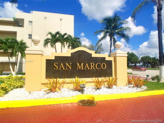 Front view of a sign for San Marco with a building and palm trees in the background.