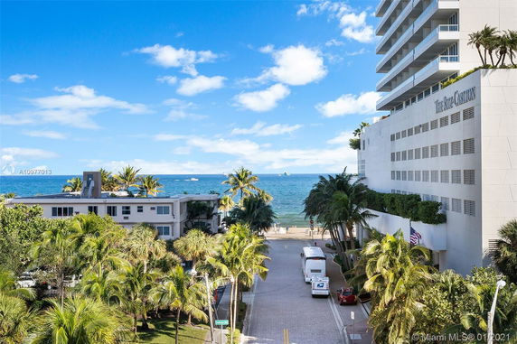 View from a building overlooking a street leading to a beach, with palm trees and a multi-story hotel on the right.