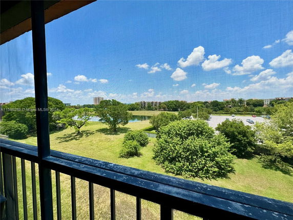 View from a building balcony overlooking grassy landscape and parking area with trees.