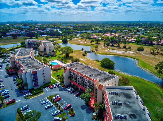 Aerial view of apartment buildings with parking lots, surrounded by greenery and a body of water.