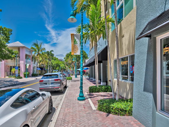 Street view of buildings with cars parked along the sidewalk, featuring palm trees and a clear sky.