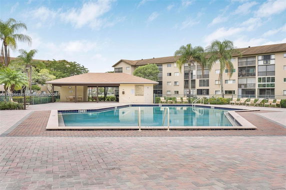 Outdoor swimming pool area near a residential building with lounge chairs and palm trees.