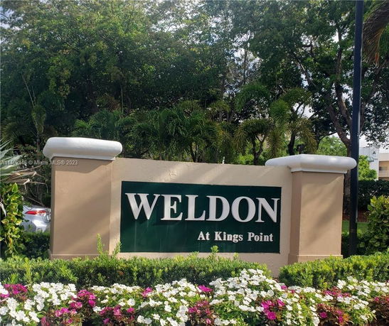 Sign for Weldon at Kings Point surrounded by trees and flowers.