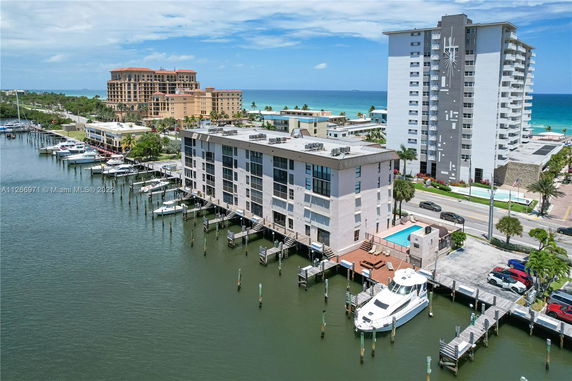 Aerial view of a multi-story waterfront building with a swimming pool, surrounded by other buildings and a marina with boats.