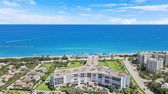 Aerial view of a large residential building near the beach with surrounding greenery and ocean in the background.