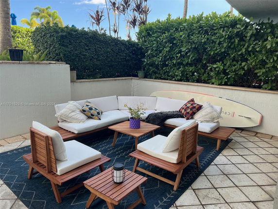 Outdoor seating area with white cushioned wooden furniture and a surfboard against the wall.