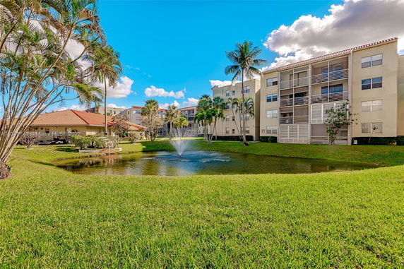 View of an apartment complex with a water fountain in the pond and palm trees.