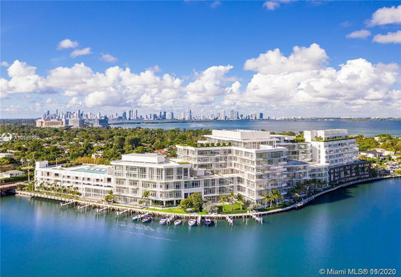 Aerial view of a waterfront multi-story residential complex with modern architecture, surrounded by water and greenery, with a city skyline in the background.
