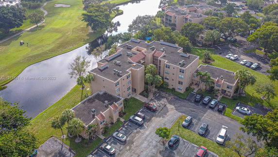 Aerial view of a large apartment complex with flat rooftops featuring air conditioning units. The building is surrounded by a parking area and greenery, with a water body and golf course nearby.