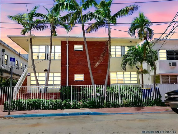 Front view of a two-story building with a brick facade and large windows, surrounded by palm trees.