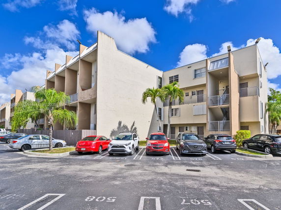 Exterior view of a multi-story residential building with adjacent parking spaces filled with cars.