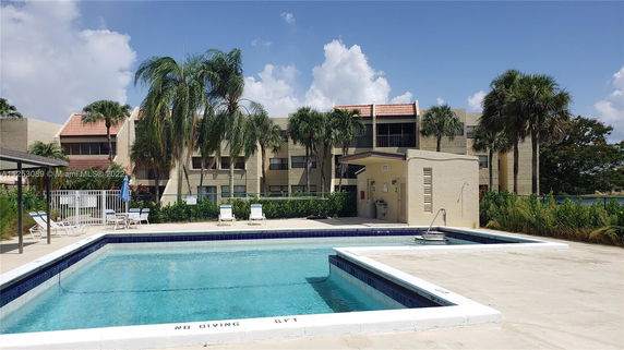 Outdoor swimming pool area with surrounding palm trees and a multi-story building in the background.
