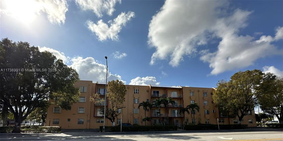 Front view of a multi-story residential building with balconies and trees surrounding it.