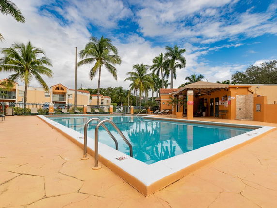 Outdoor swimming pool area with palm trees and surrounding buildings.