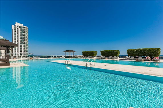 A large outdoor swimming pool with clear blue water, surrounded by deck chairs and green shrubs, adjacent to a tall residential building.