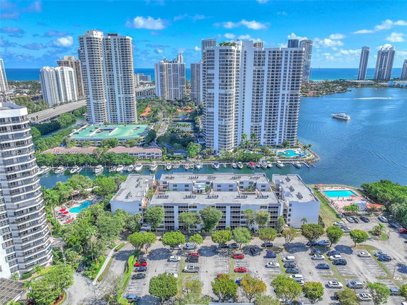 View of multiple high-rise buildings near a body of water with surrounding greenery and a parking area.
