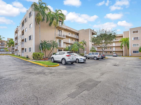 Front view of a multi-story apartment building with balconies and adjacent parking area.