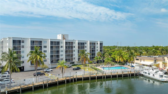 View of a multi-story apartment building adjacent to water, with a visible parking area and swimming pool.
