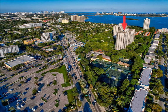 Aerial view of a cityscape with residential and commercial buildings, roads, and green areas near a body of water.